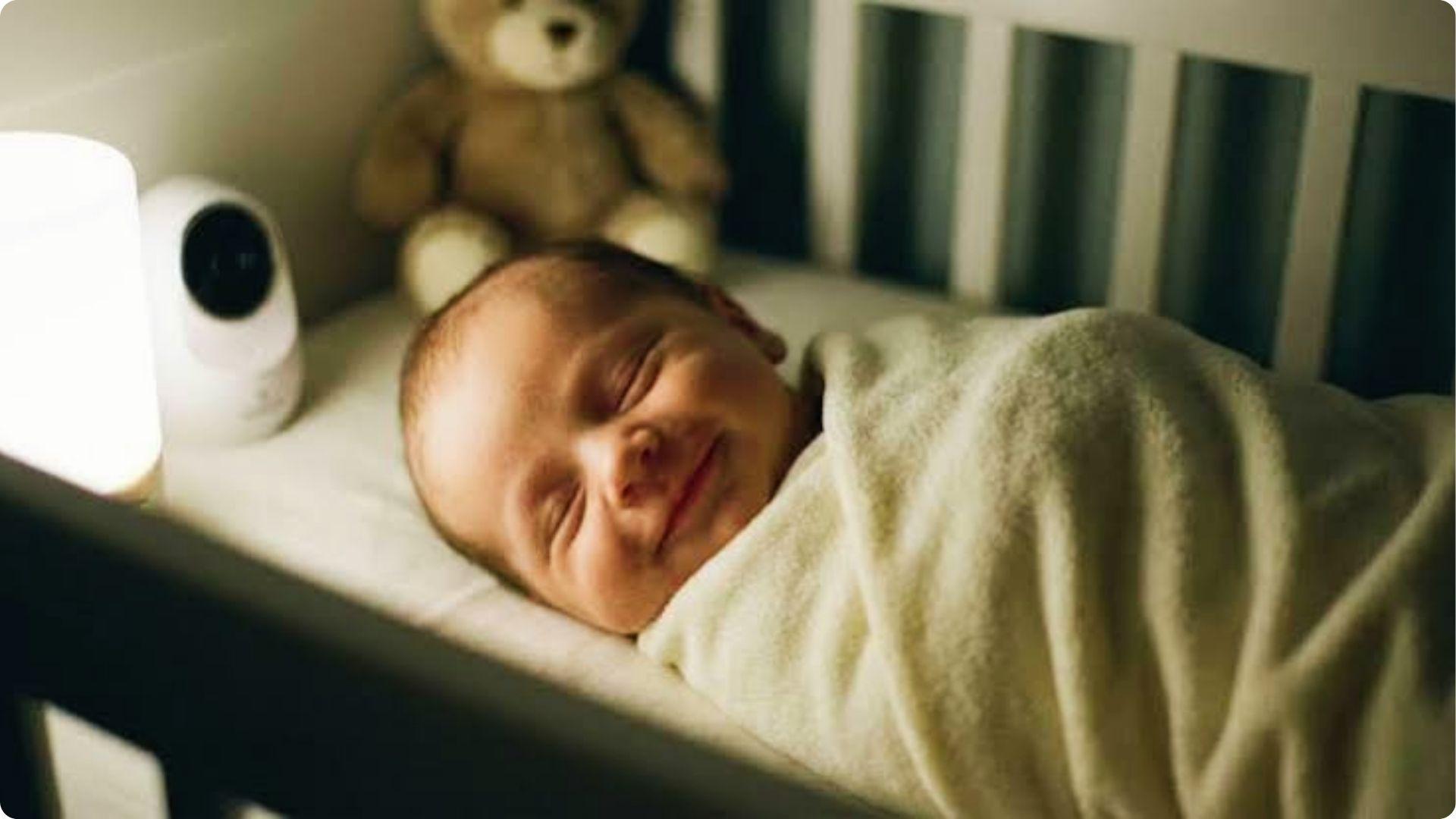 Swaddled newborn smiling in crib near teddy and monitor, showing why babies smile in sleep during dream stage