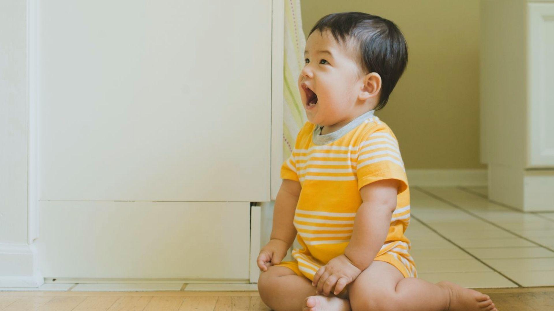 Joyful baby in yellow striped onesie sitting on wooden floor laughing happily indoors