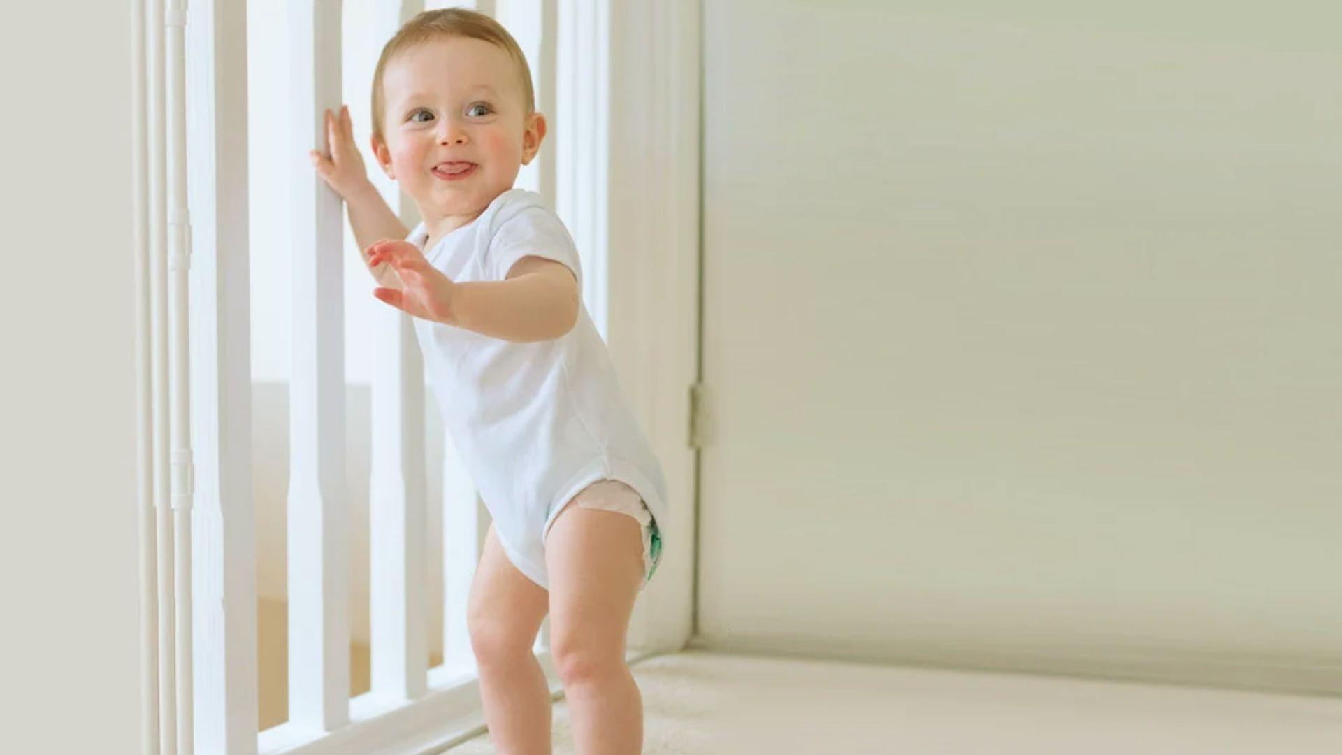 Happy baby in white onesie standing and holding white safety gate indoors learning to walk