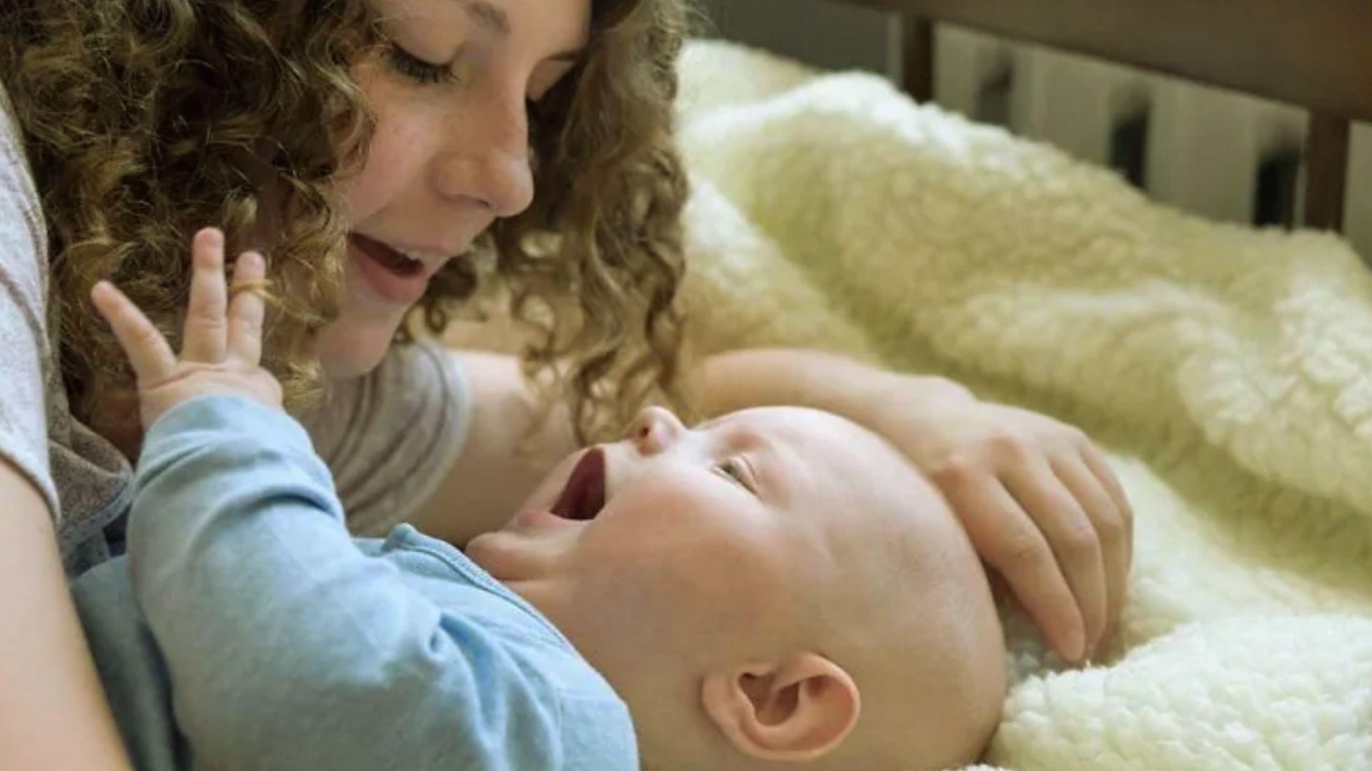 Curly-haired mother playing joyfully with laughing baby in blue outfit on cozy blanket