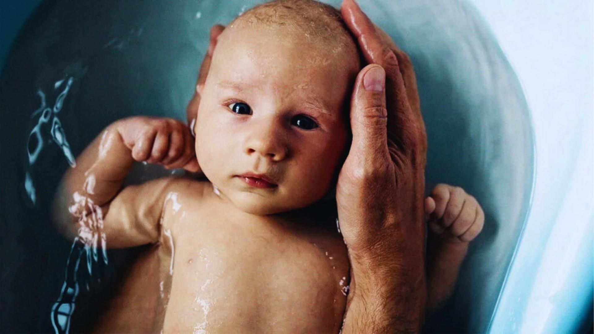 Newborn baby bathing in tub supported by adult hands during gentle bath time care today.