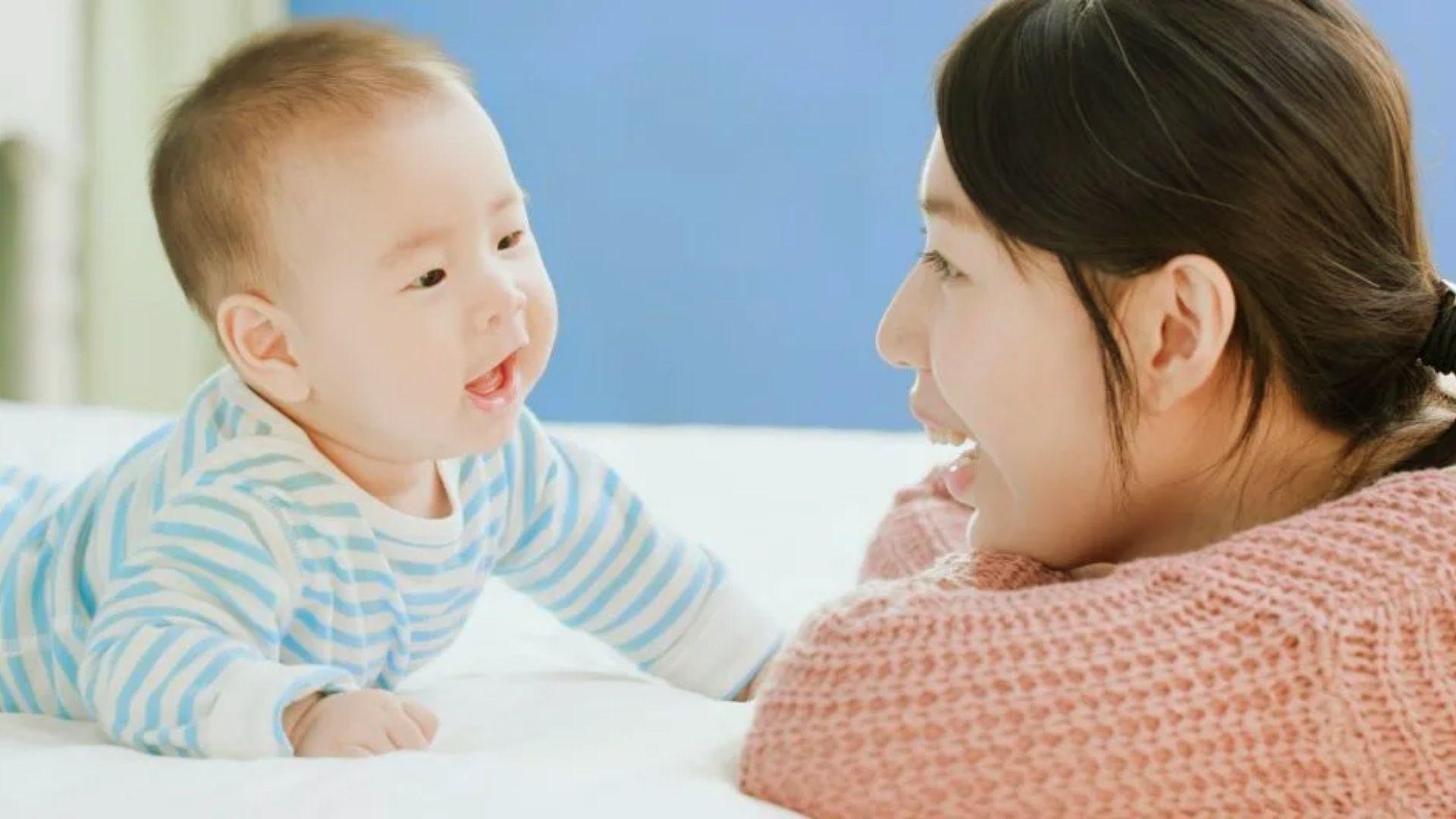 Happy mother in pink sweater smiling at baby in blue striped onesie during tummy time on bed