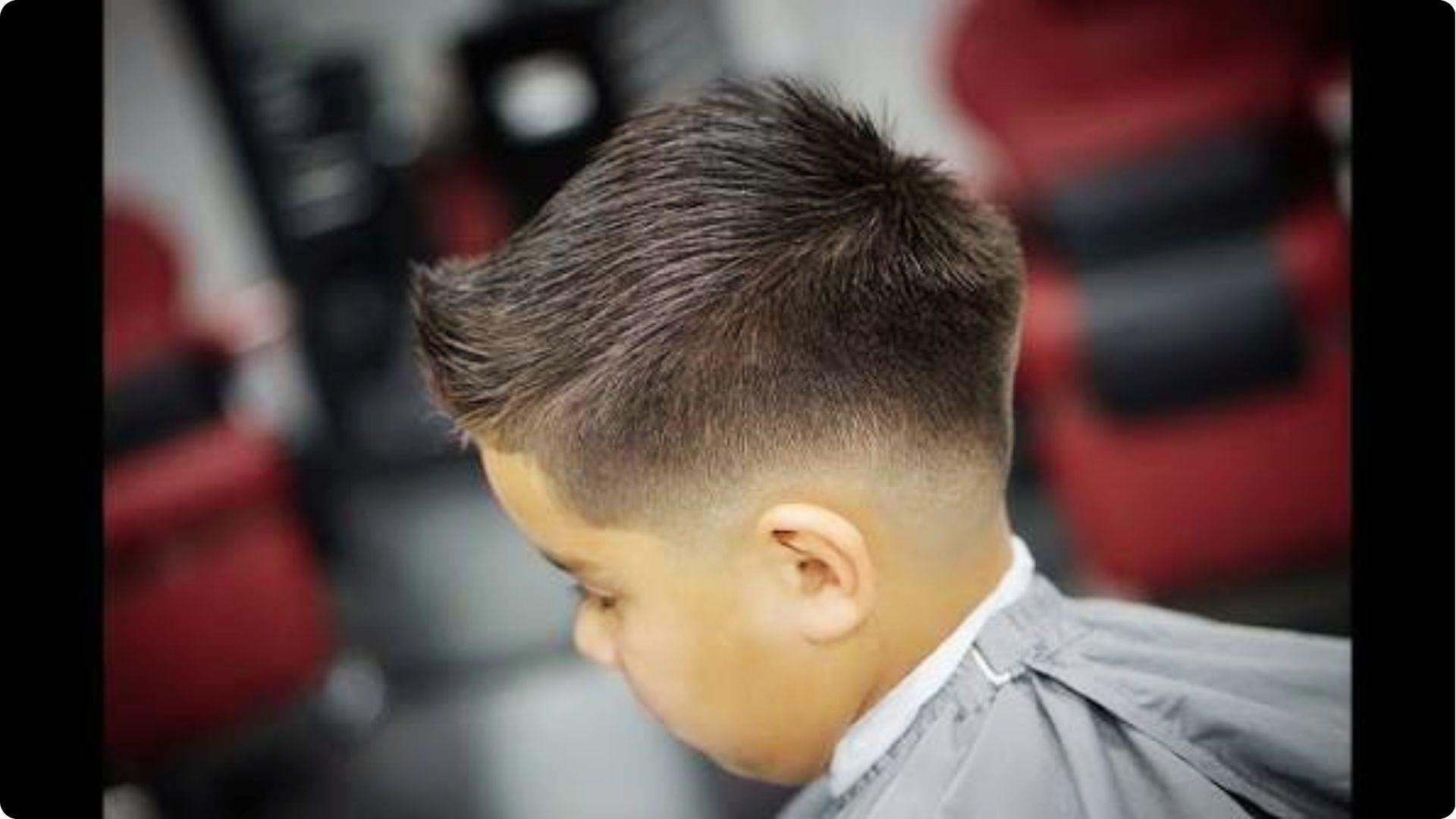 Toddler boy in barber cape with high fade and spiky quiff pompadour, side view, trendy baby haircut.