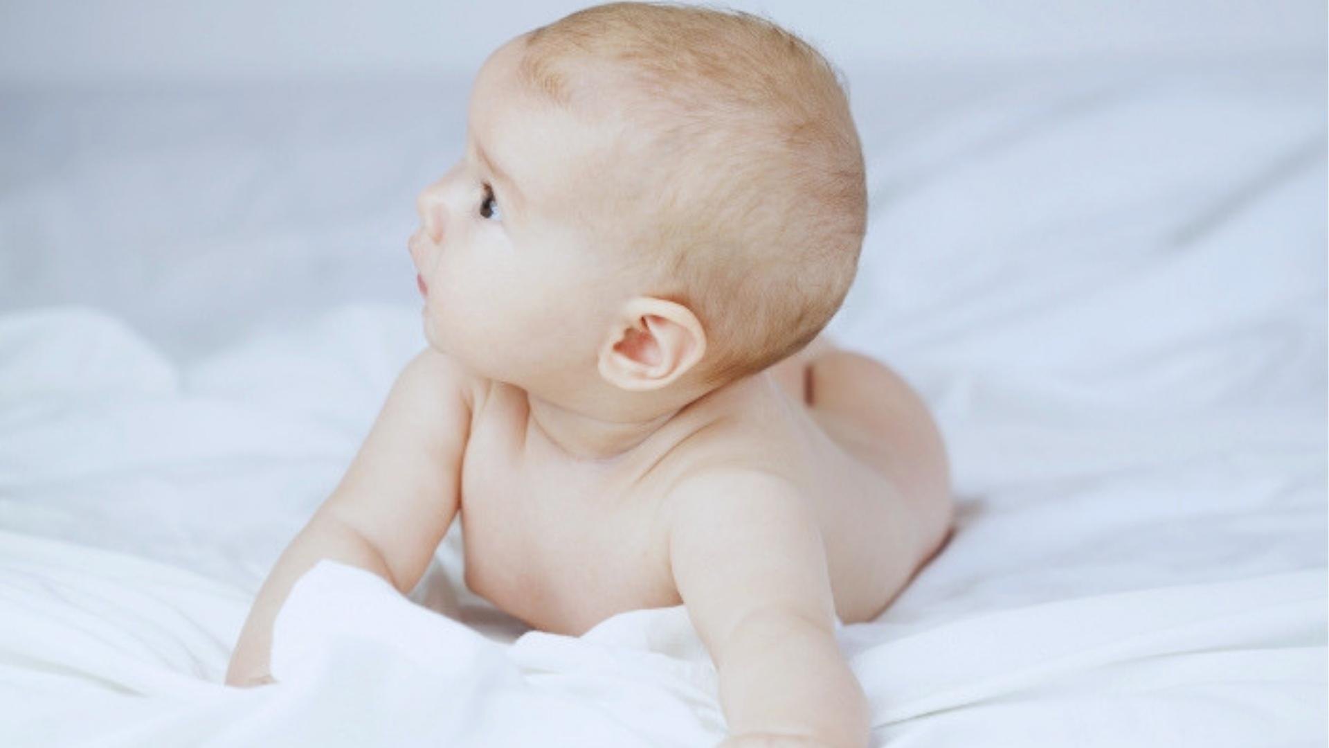 Baby lying on white bedsheet looking sideways during tummy time developmental milestone moment today happily.
