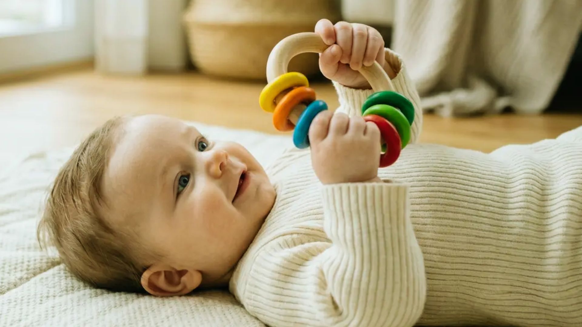 Happy baby lying on blanket holding colorful wooden ring rattle toy with both hands