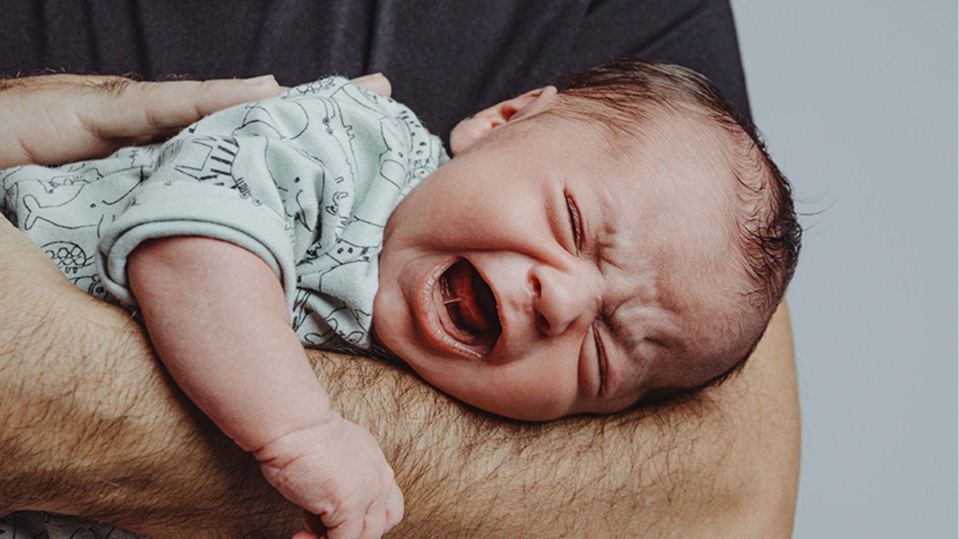 Baby cries loudly while lying on father’s arm, signaling hunger or discomfort needing attention.