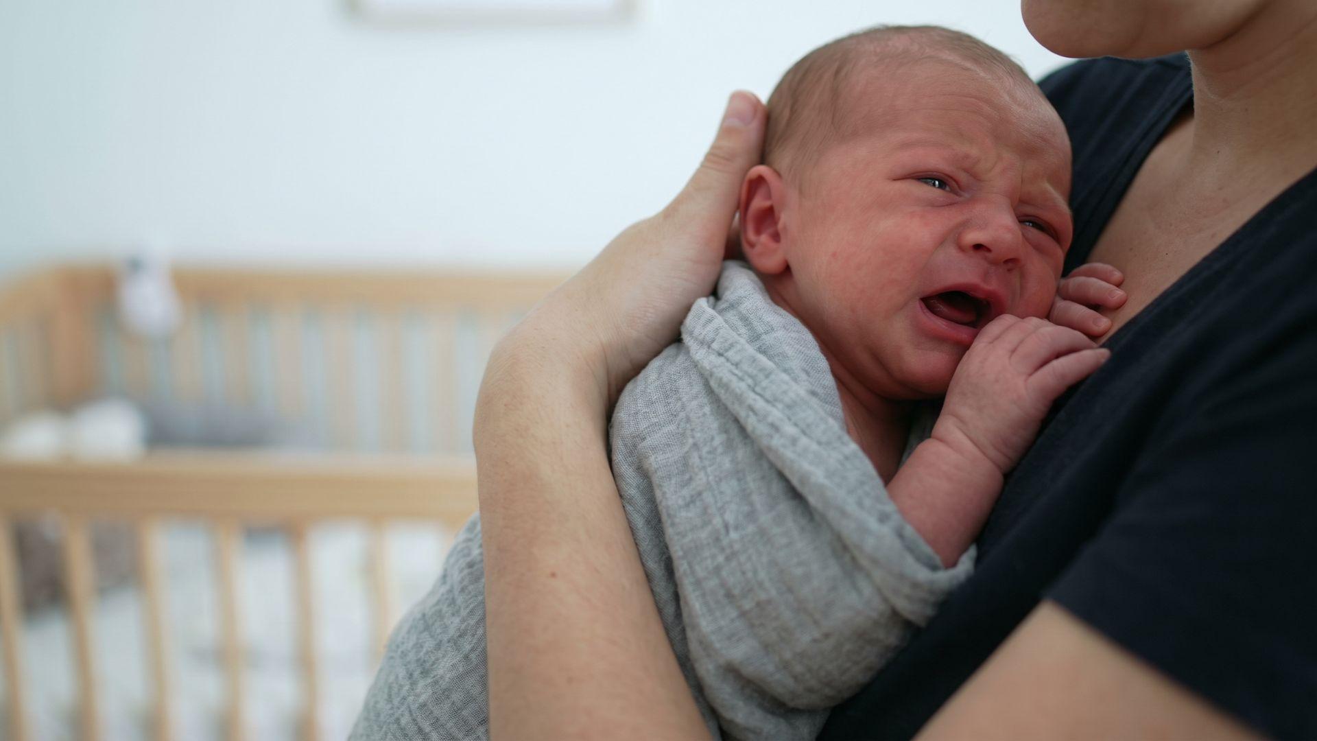 Parent holding a crying newborn close to their chest, with the baby’s mouth open and hands near the chest.