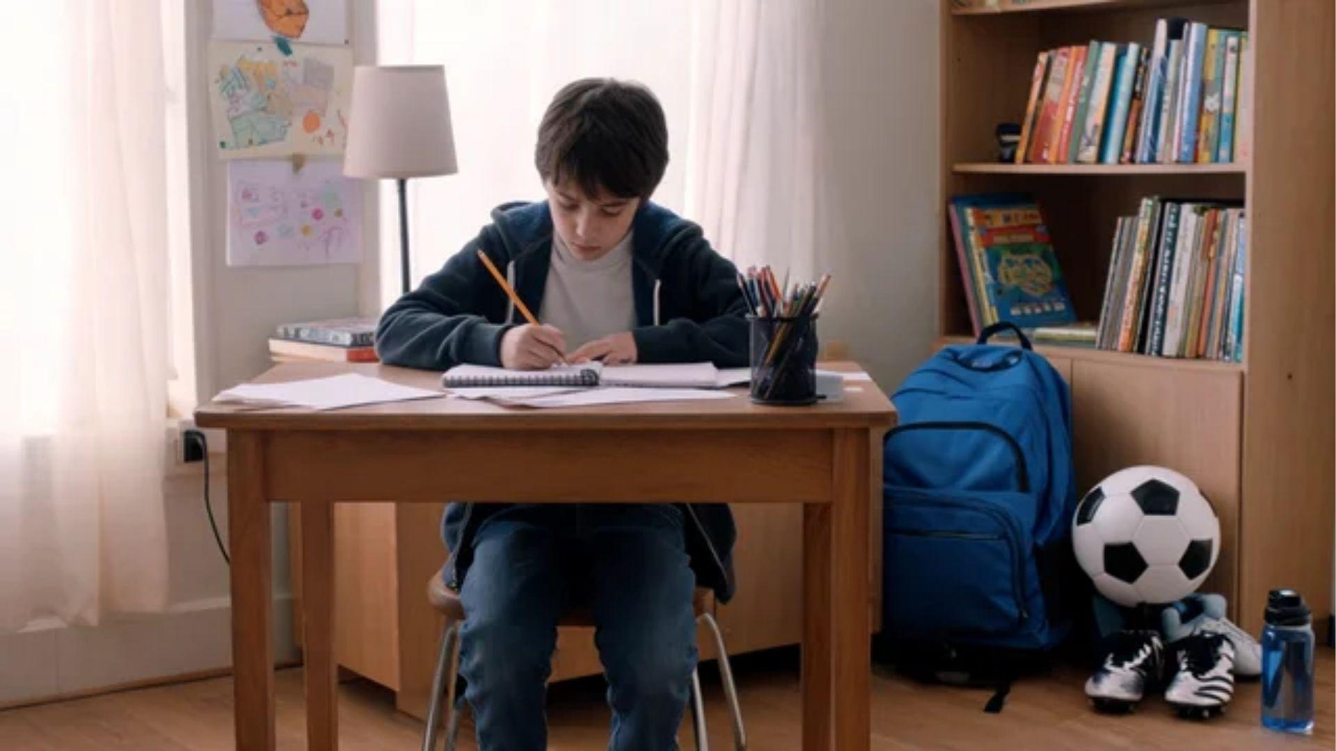 School-aged child writing in a notebook beside a schoolbag and sports equipment