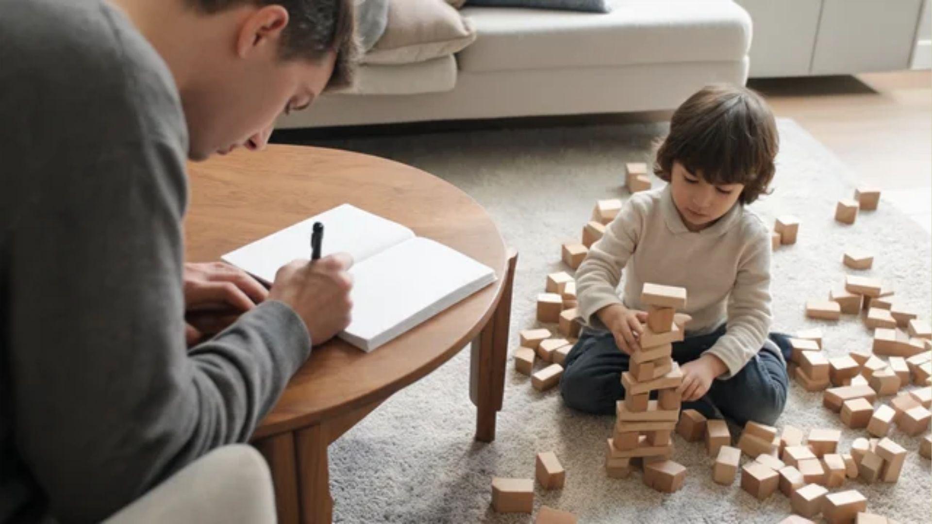Parent writing in a notebook while a young child builds with wooden blocks nearby