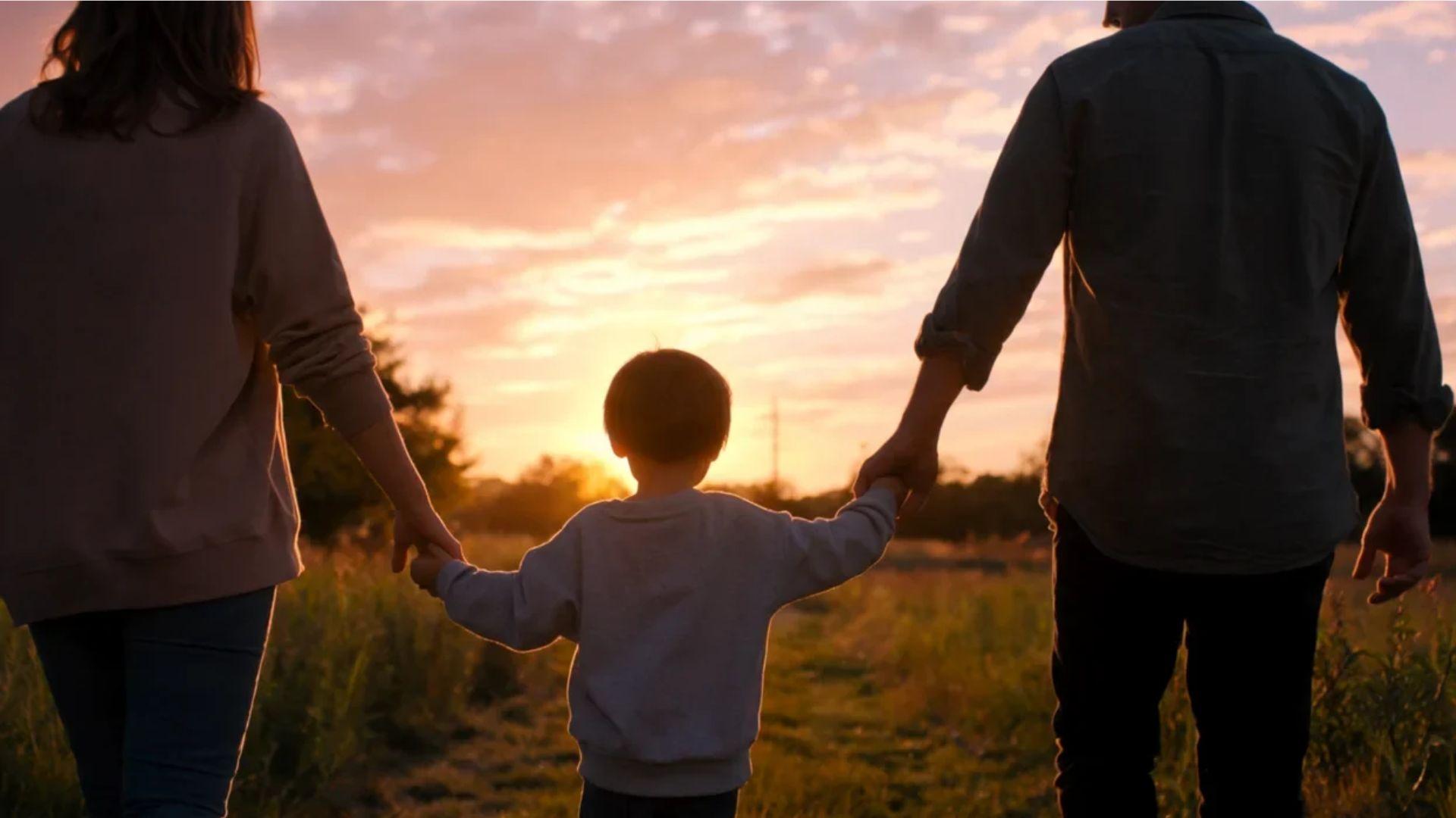 Child holding parents’ hands walking outdoors at sunset from behind.