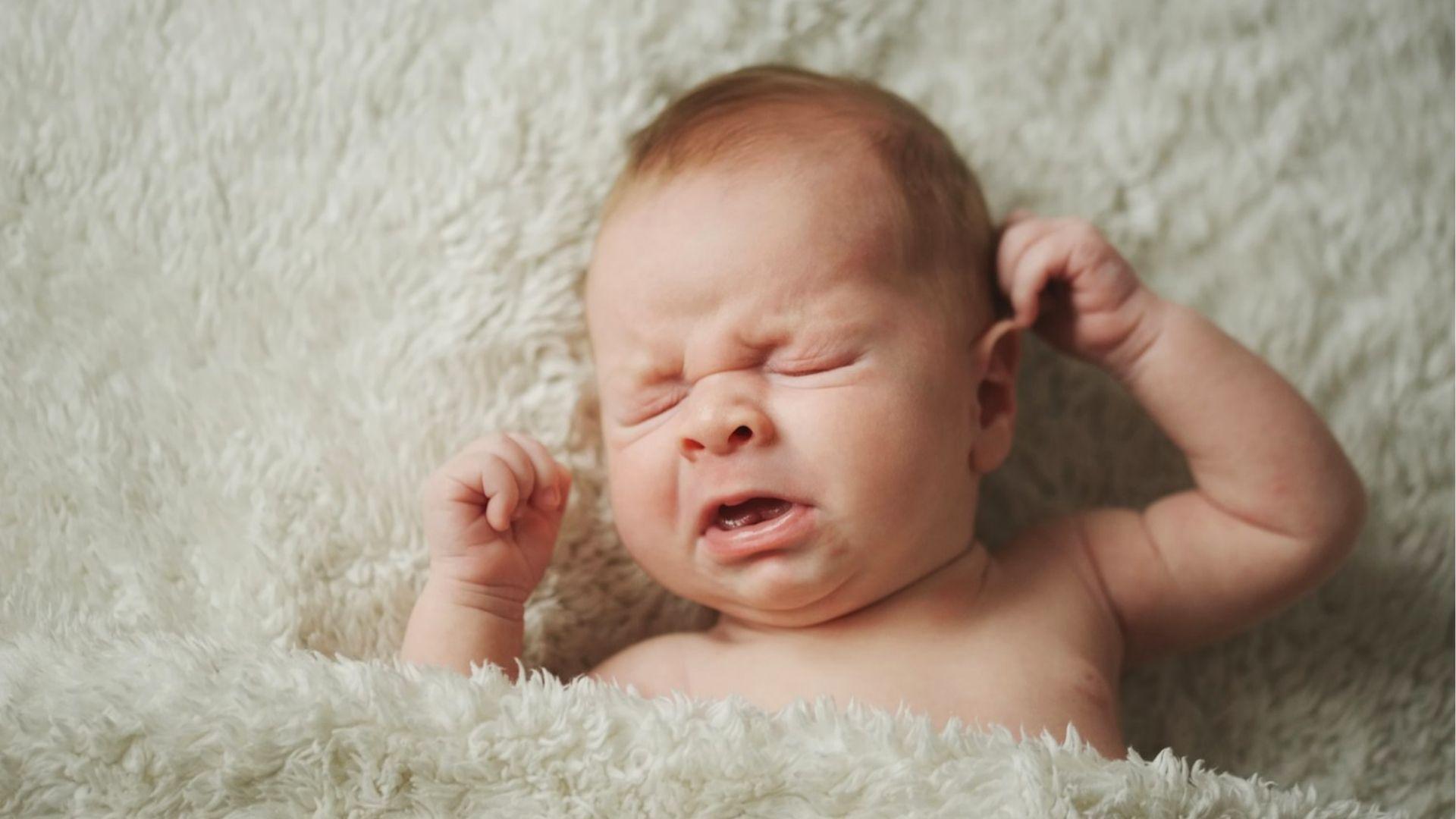 A newborn baby crying with fists raised while lying on a soft white blanket.