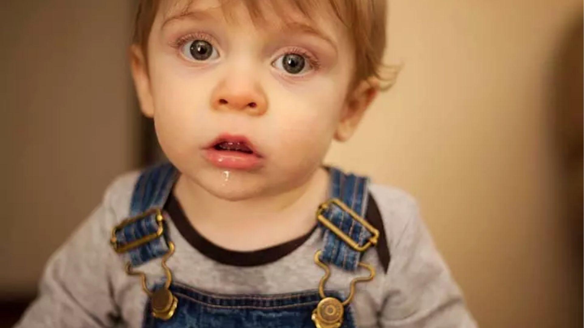 Toddler in overalls with drool on chin close-up, showing excessive drooling.