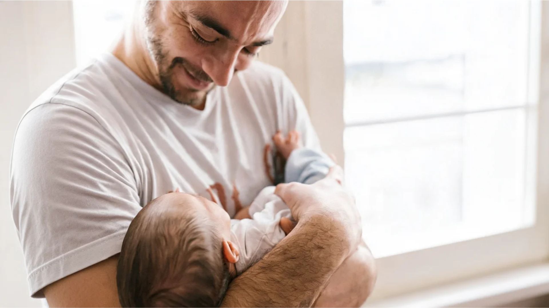 Father smiling while holding newborn baby in arms near window