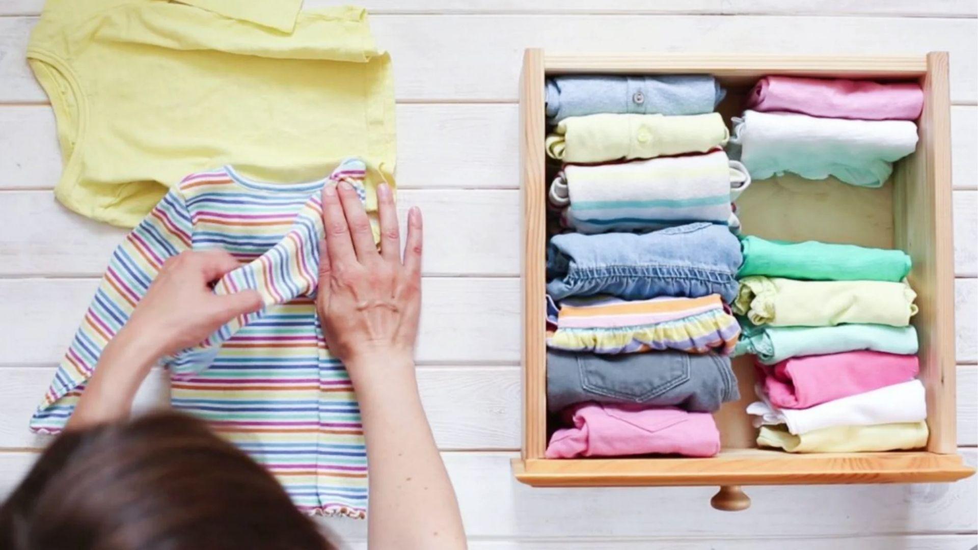 Overhead hands folding striped baby shirt on white wood beside drawer.