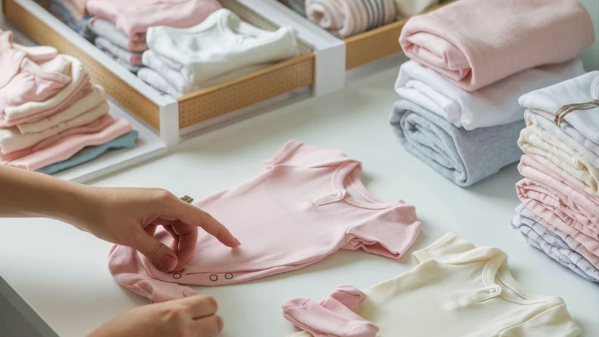 Hand folding pink baby bodysuit on white table surrounded by stacked folded pastel onesies.
