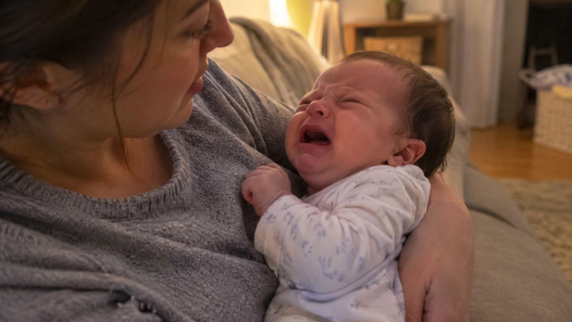 Parent cradling a crying infant in their arms at home during the evening.
