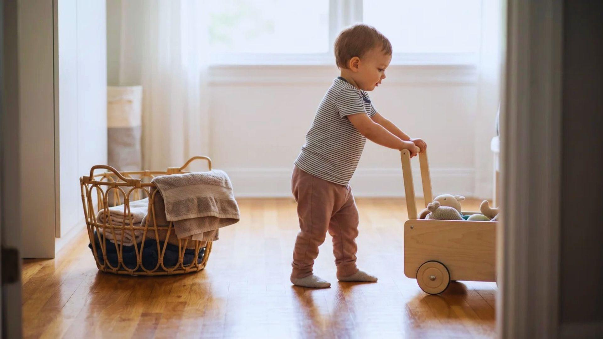 Toddler pushing a lightweight wagon across a clear home floor beside a laundry basket.