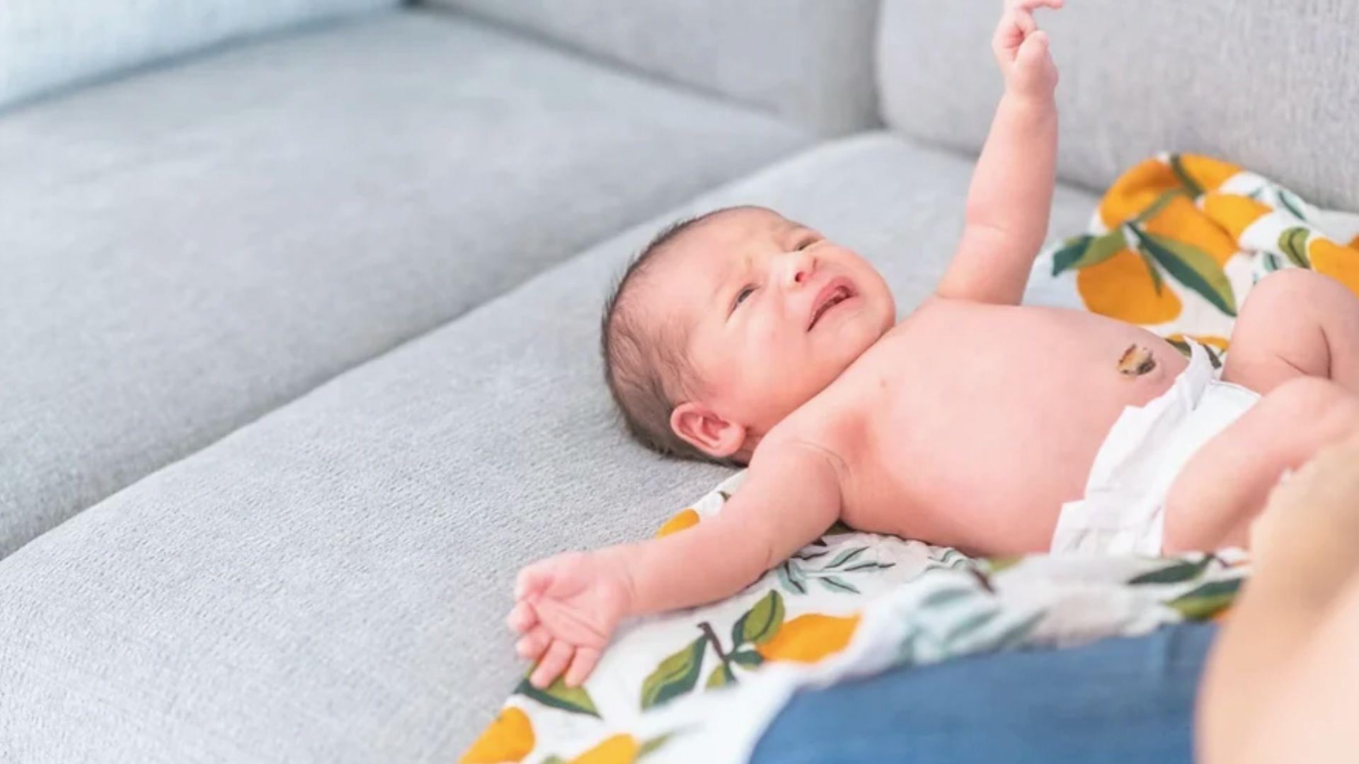 Newborn baby with umbilical cord stump lying on a gray sofa, one arm raised upward.