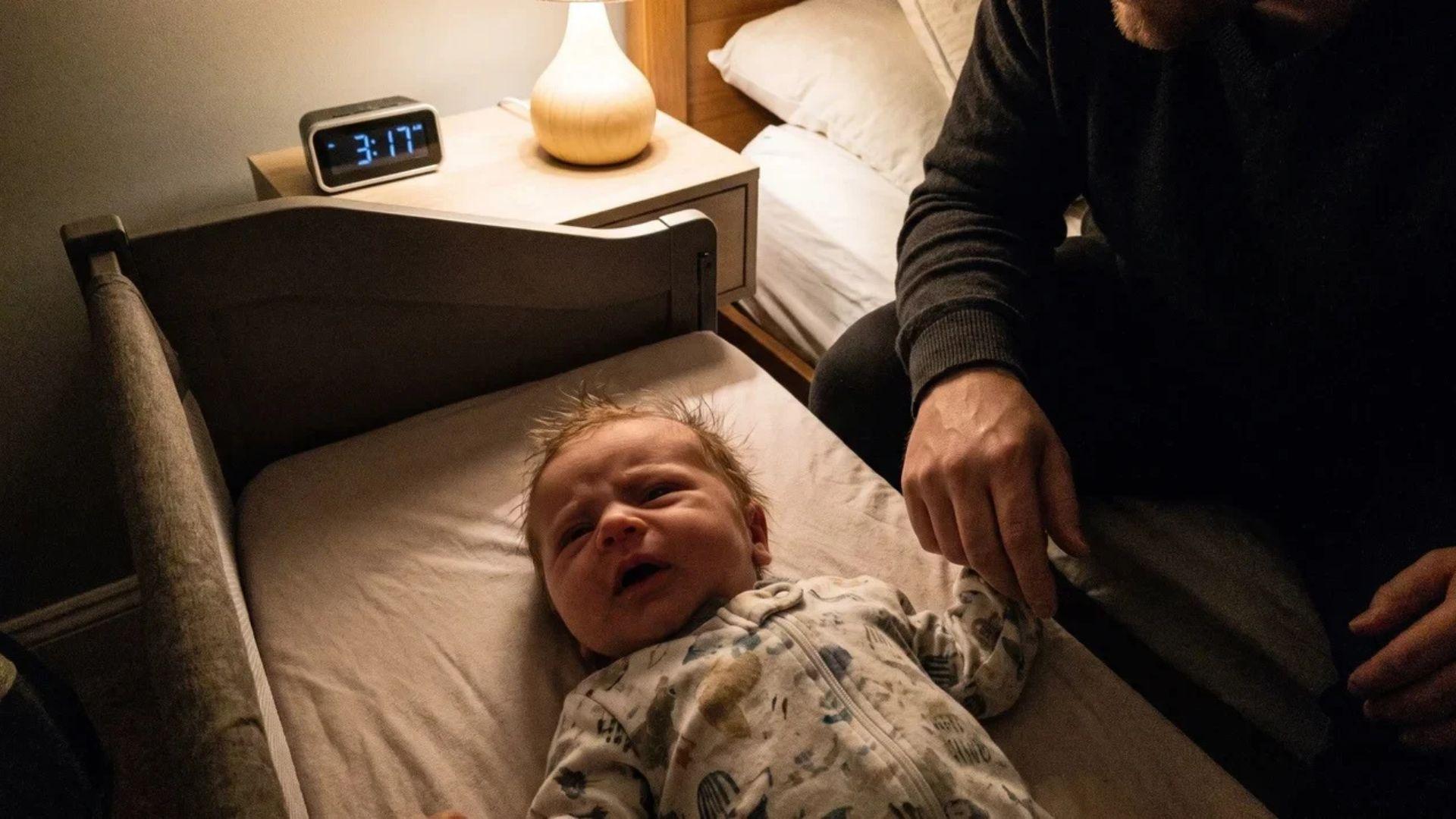 Newborn beginning to fuss in a bare bedside crib while a parent sits nearby in a dim bedroom.