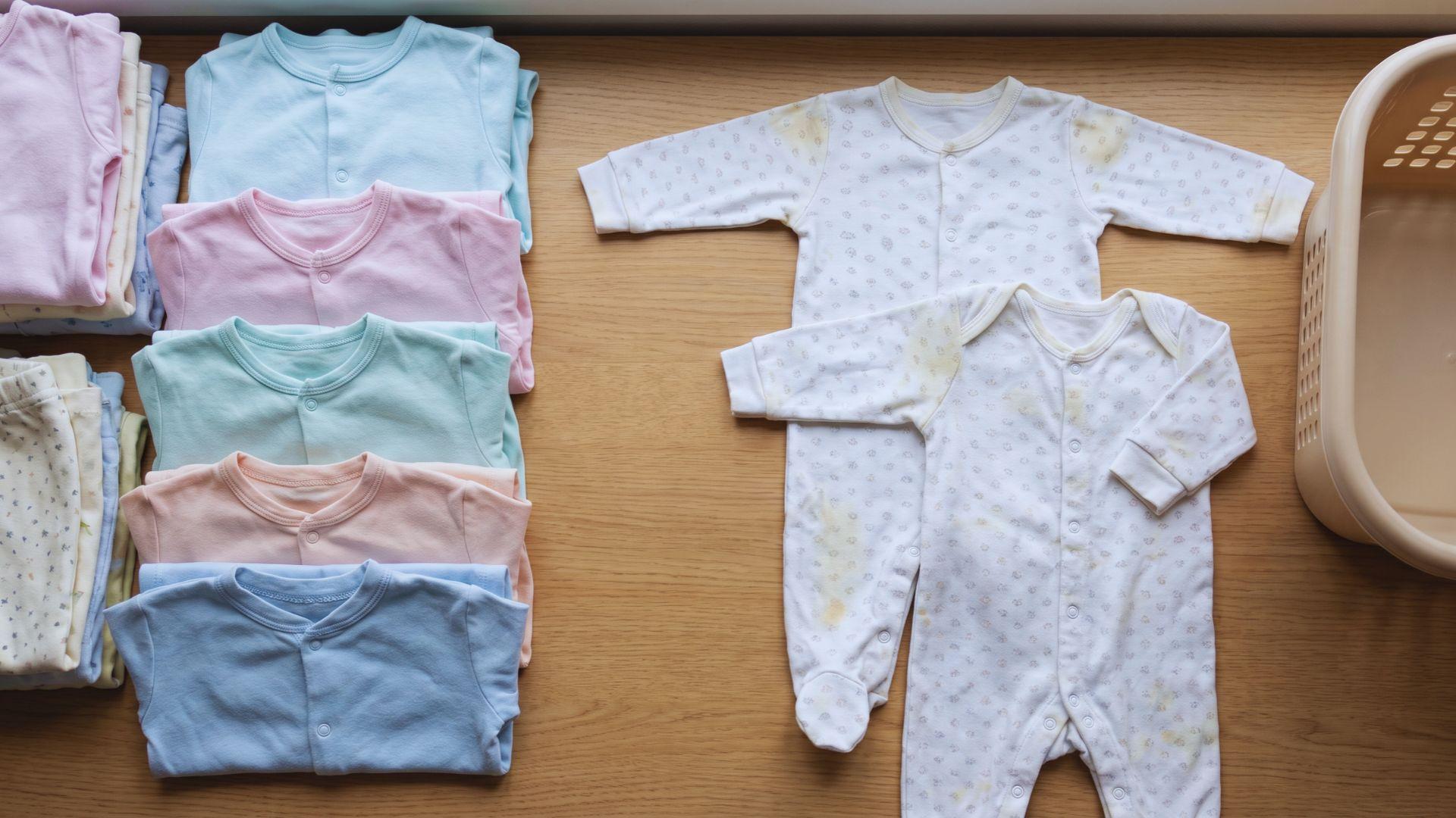 Clean baby sleepers sit beside two sleepers with small stains on a laundry surface.