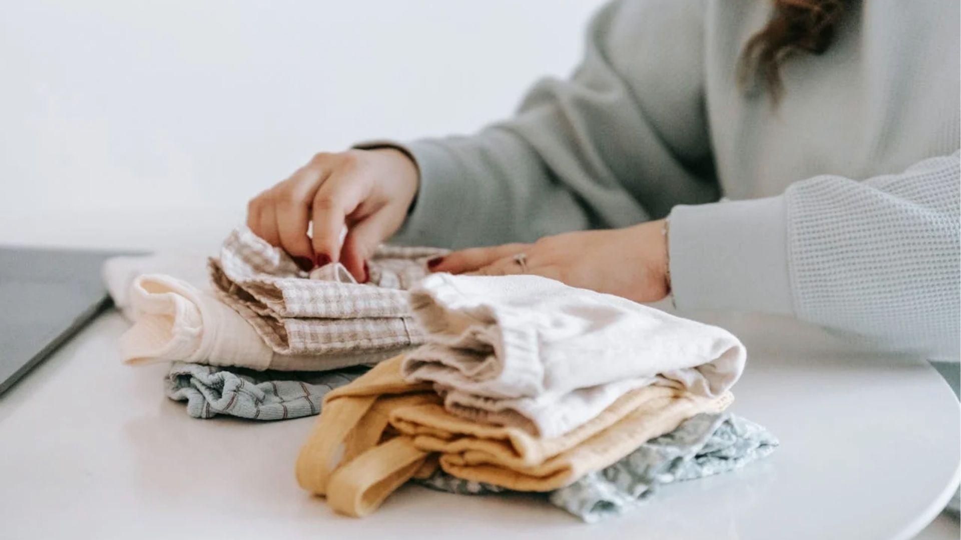 Woman’s hands folding neutral baby clothes on white table.