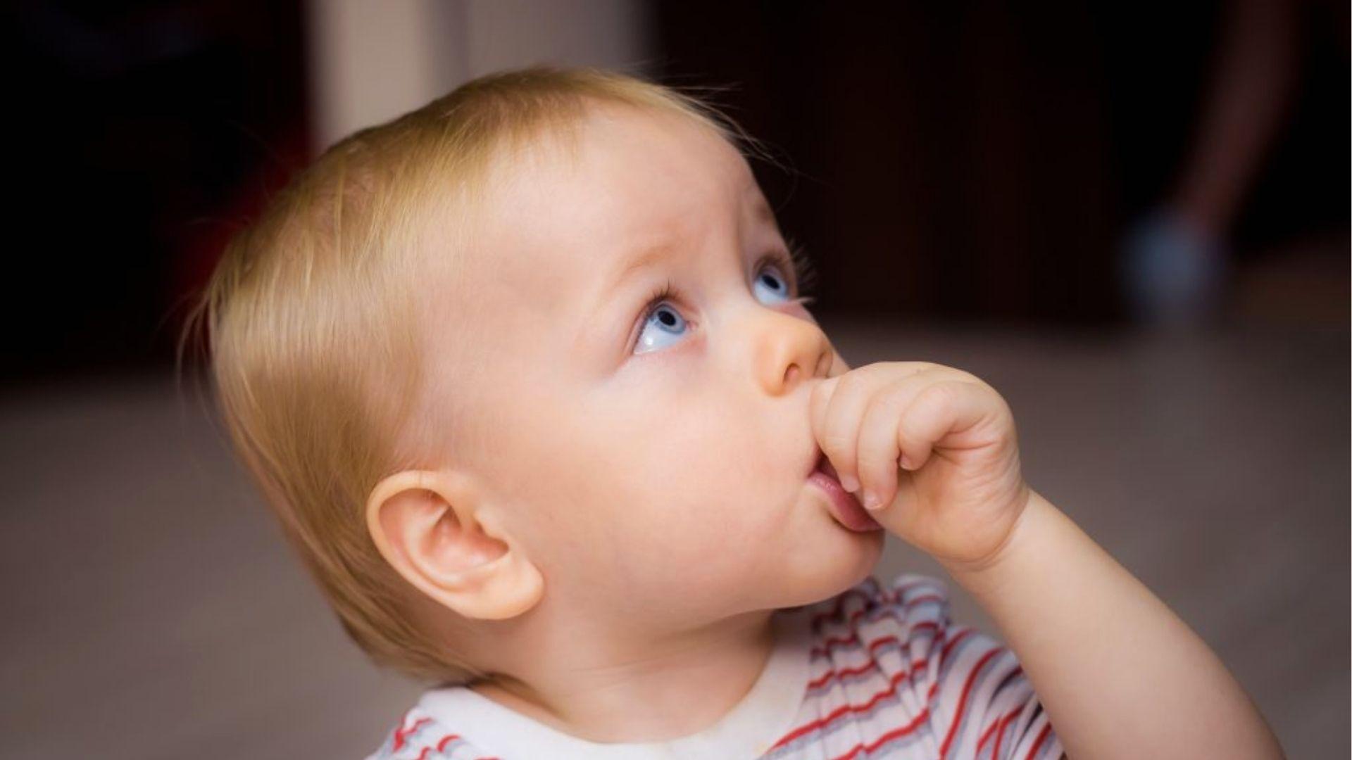 A blonde toddler sucking thumb while looking up.