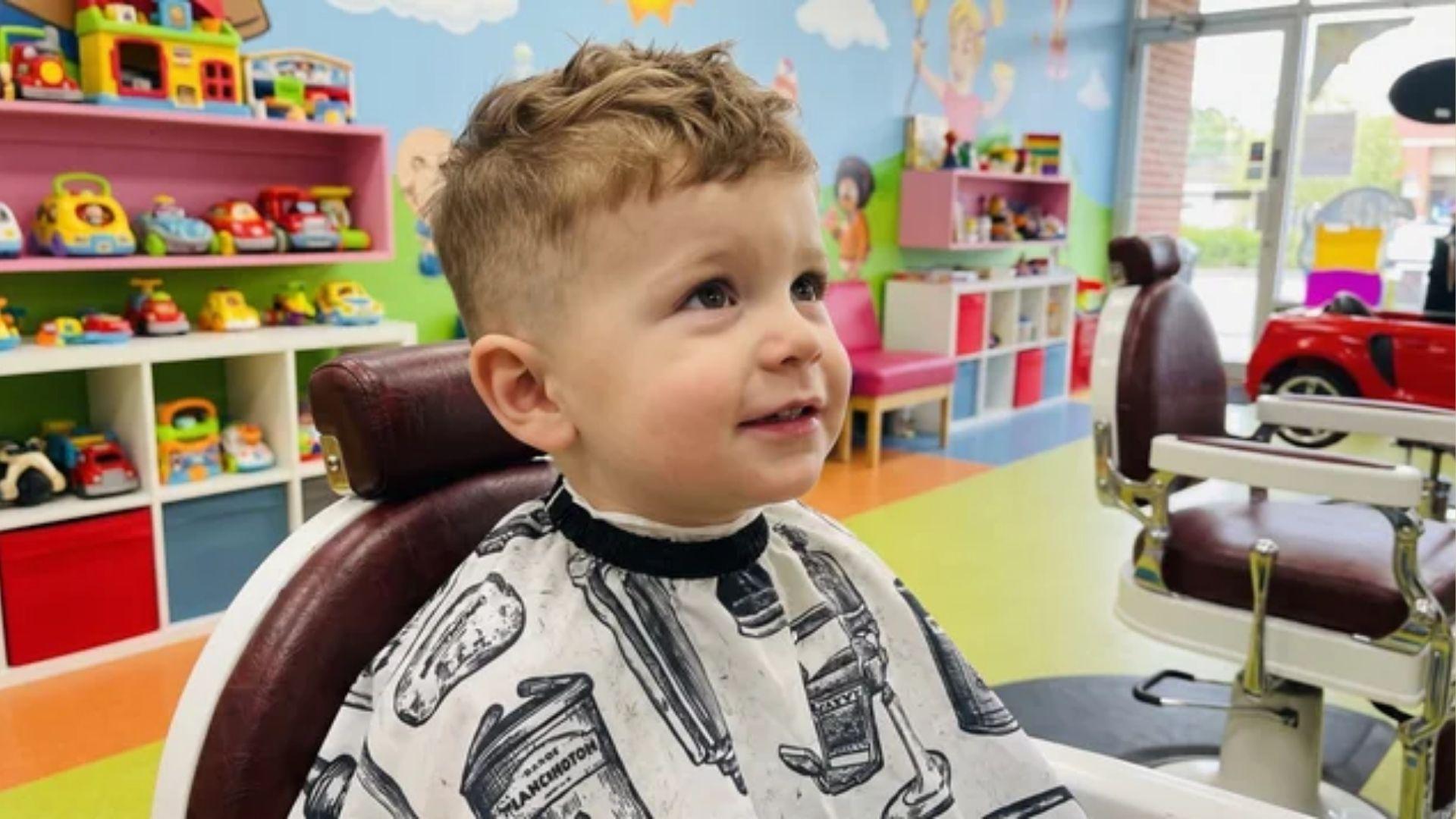 Toddler boy taper fade haircut with a short textured crop, wearing a barber cape in a kids’ barber chair