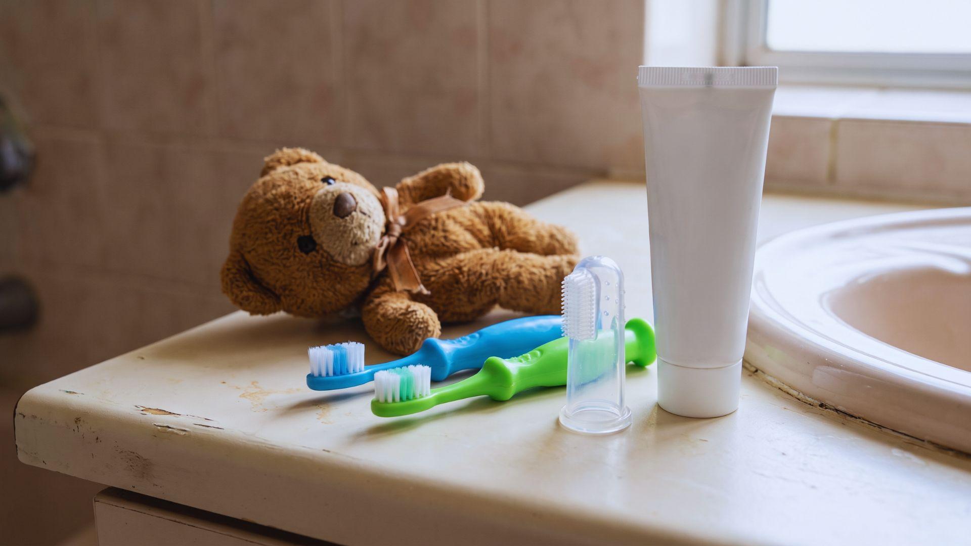 Silicone finger toothbrush, children's toothbrushes, unflavored toothpaste, and stuffed animal on a bathroom counter.