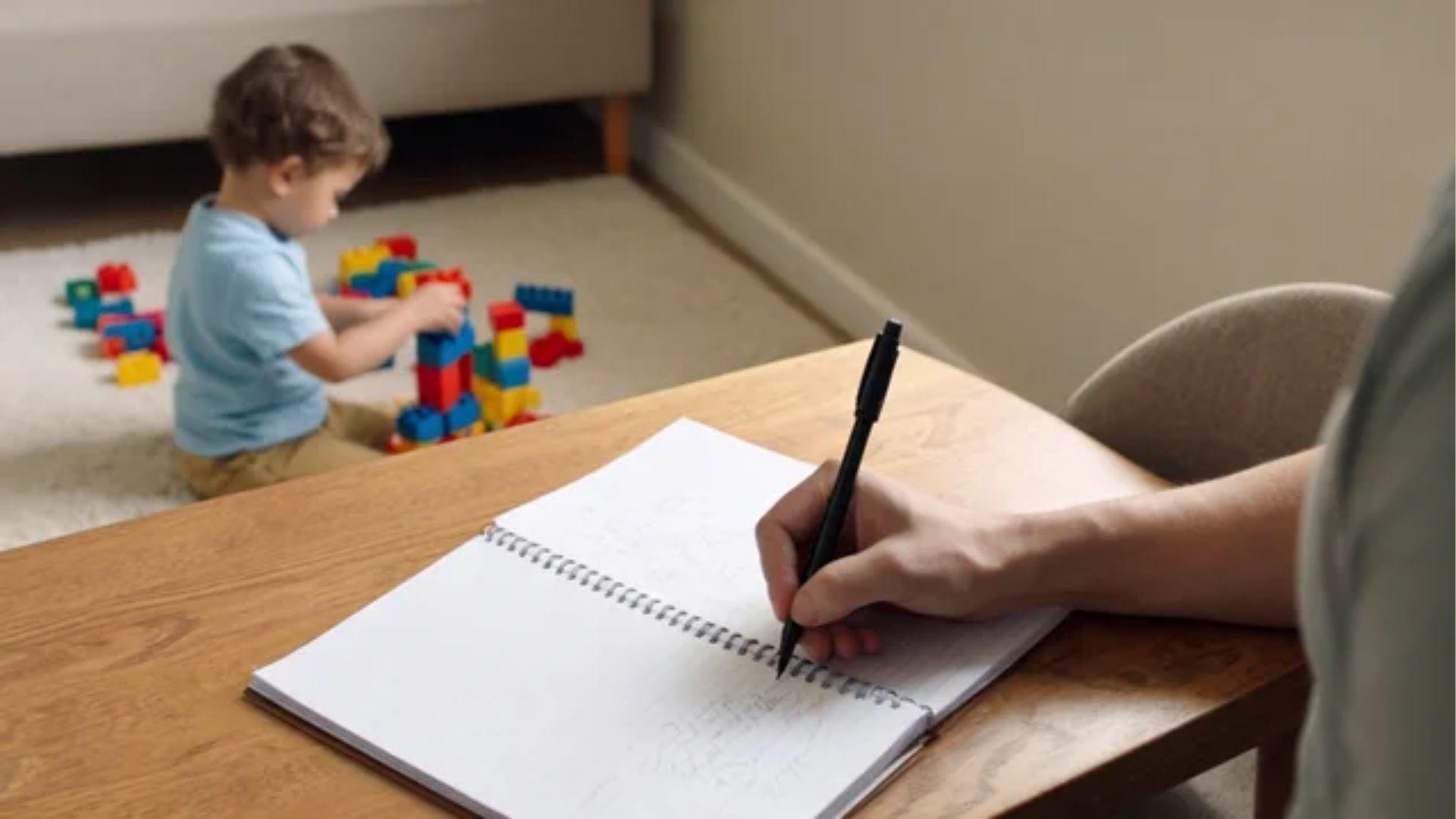 Parent recording observations in a notebook while a child plays with blocks nearby