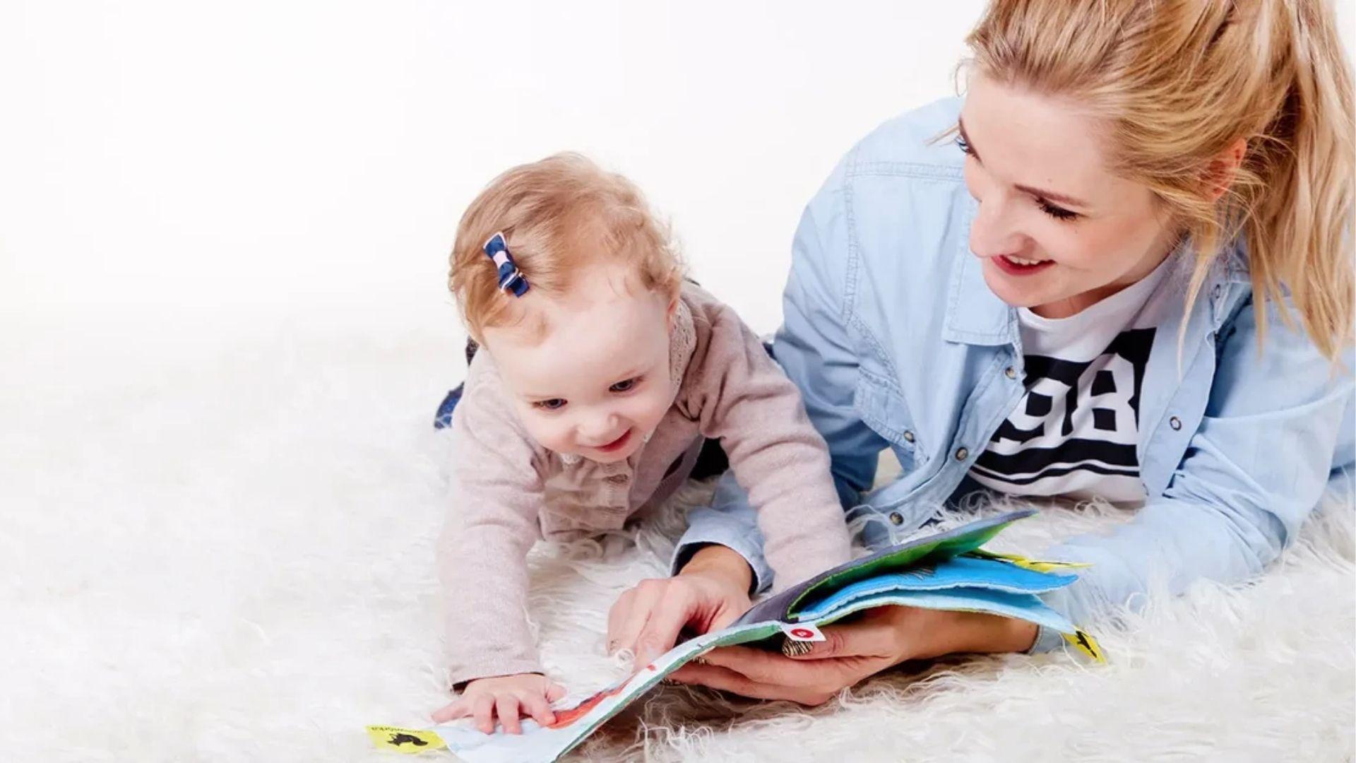Mom and baby girl on white rug reading a colorful book.