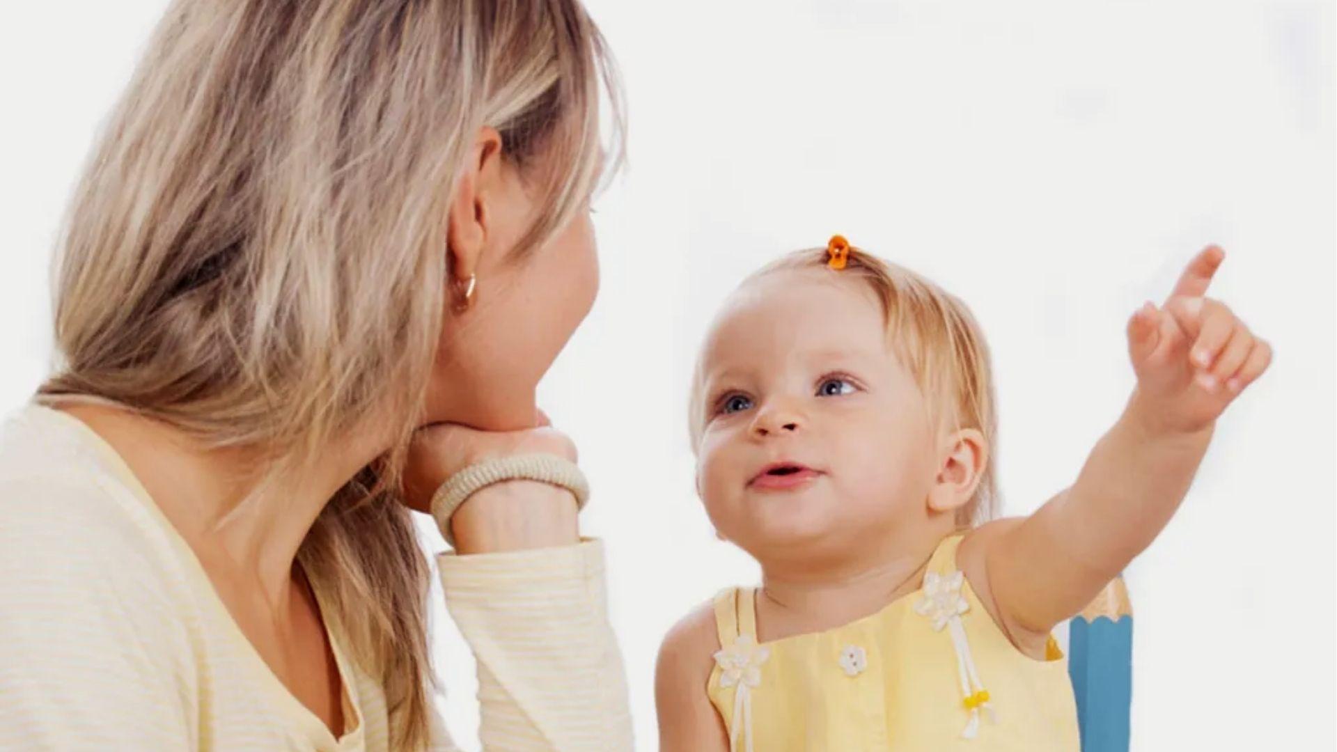 Mom listening as toddler in yellow dress points and babbles.